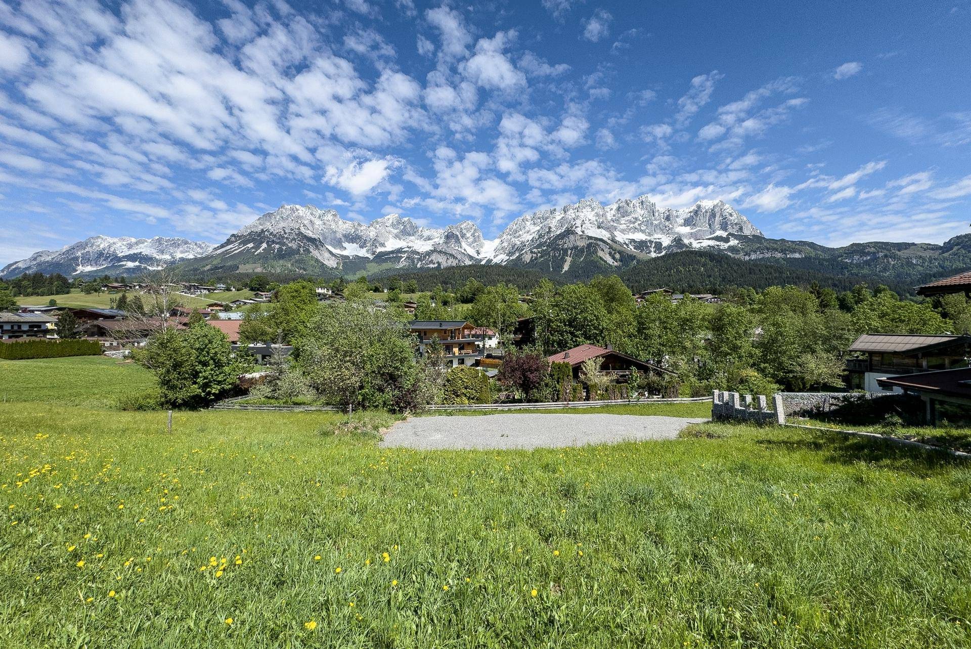 Panoramablick auf eine weite grüne Wiese mit majestätischen schneebedeckten Bergen und Häusern in der Ferne.