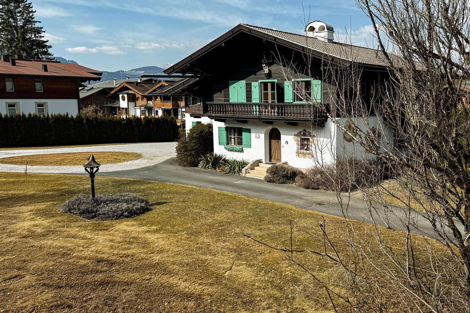 Traditionelles Alpenhaus mit grünen Fensterläden und einem kleinen Holzbalkon, umgeben von Rasen und einem Kiesweg unter blauem Himmel.