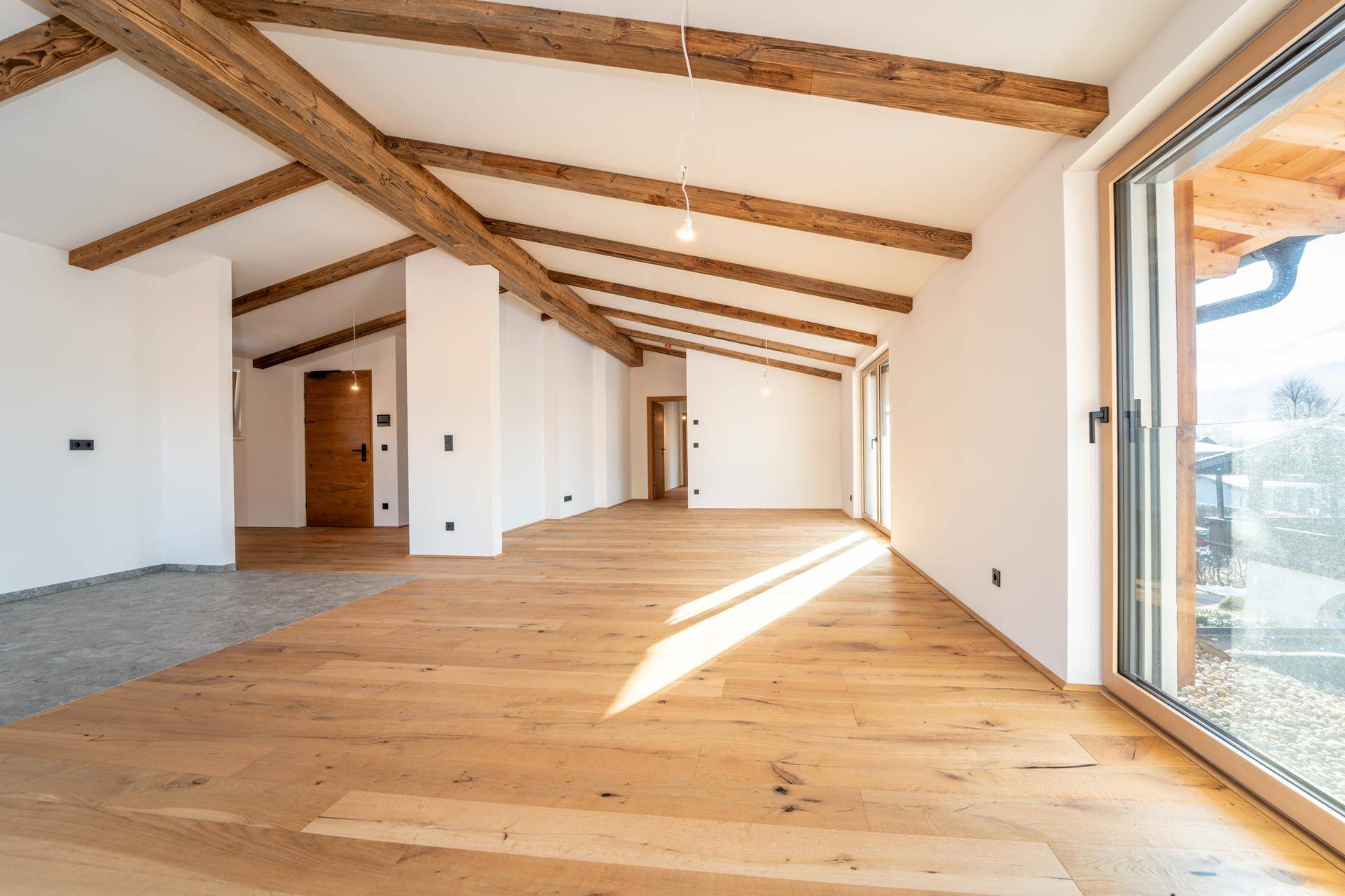 Geräumiger Wohnbereich mit sichtbaren Holzbalken, Holzboden und viel natürlichem Licht durch große Fenster.