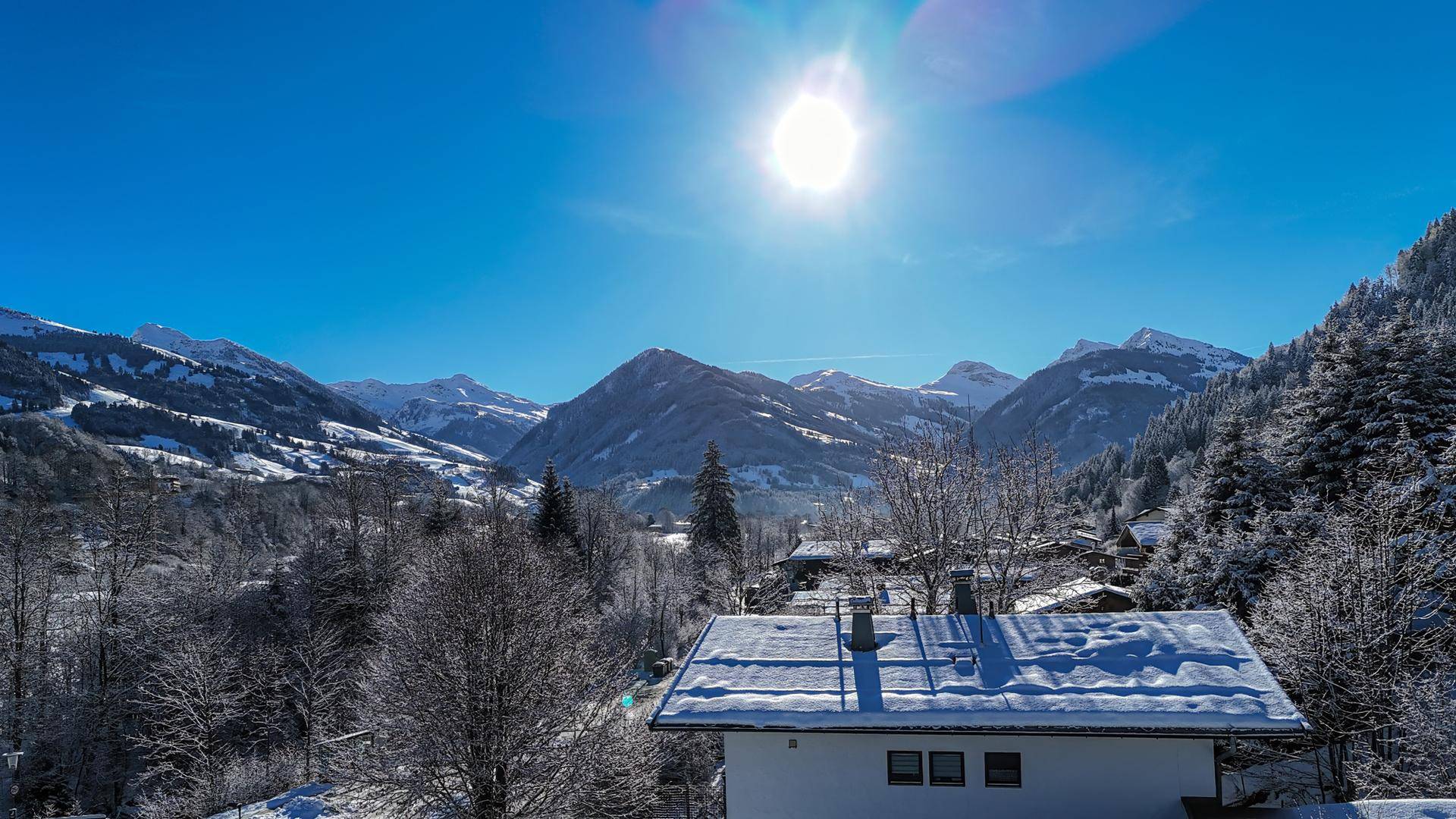 Sonniger Wintertag mit Blick auf schneebedeckte Berge und ein Haus im Vordergrund.