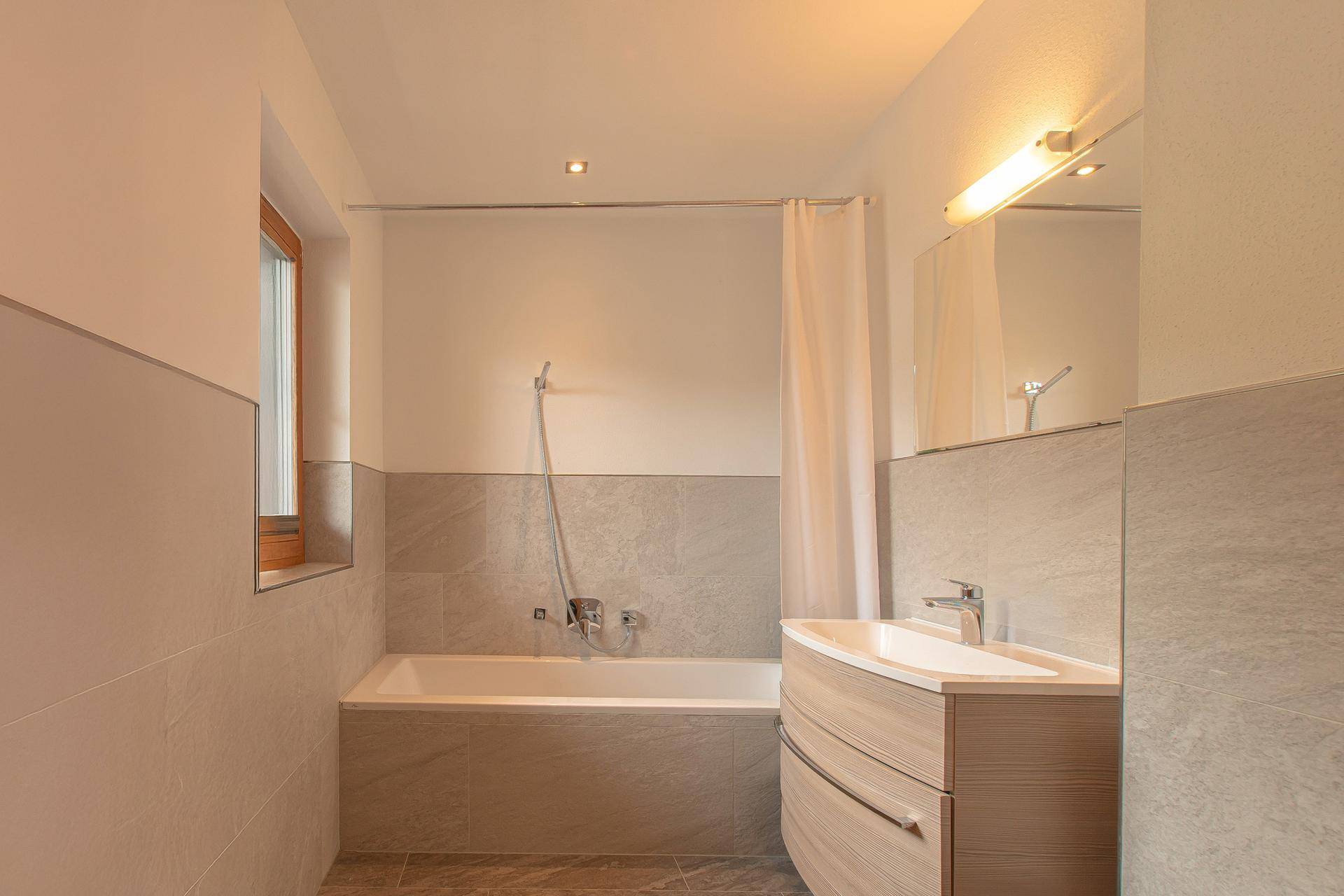 Helles Badezimmer mit Badewanne, Waschtisch und Fenster für natürliche Belüftung.