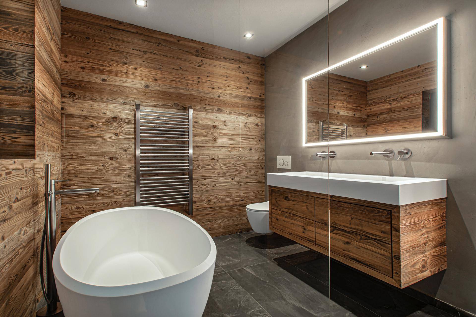 Modernes Badezimmer mit freistehender Badewanne, Holzverkleidung und beleuchtetem Spiegel.