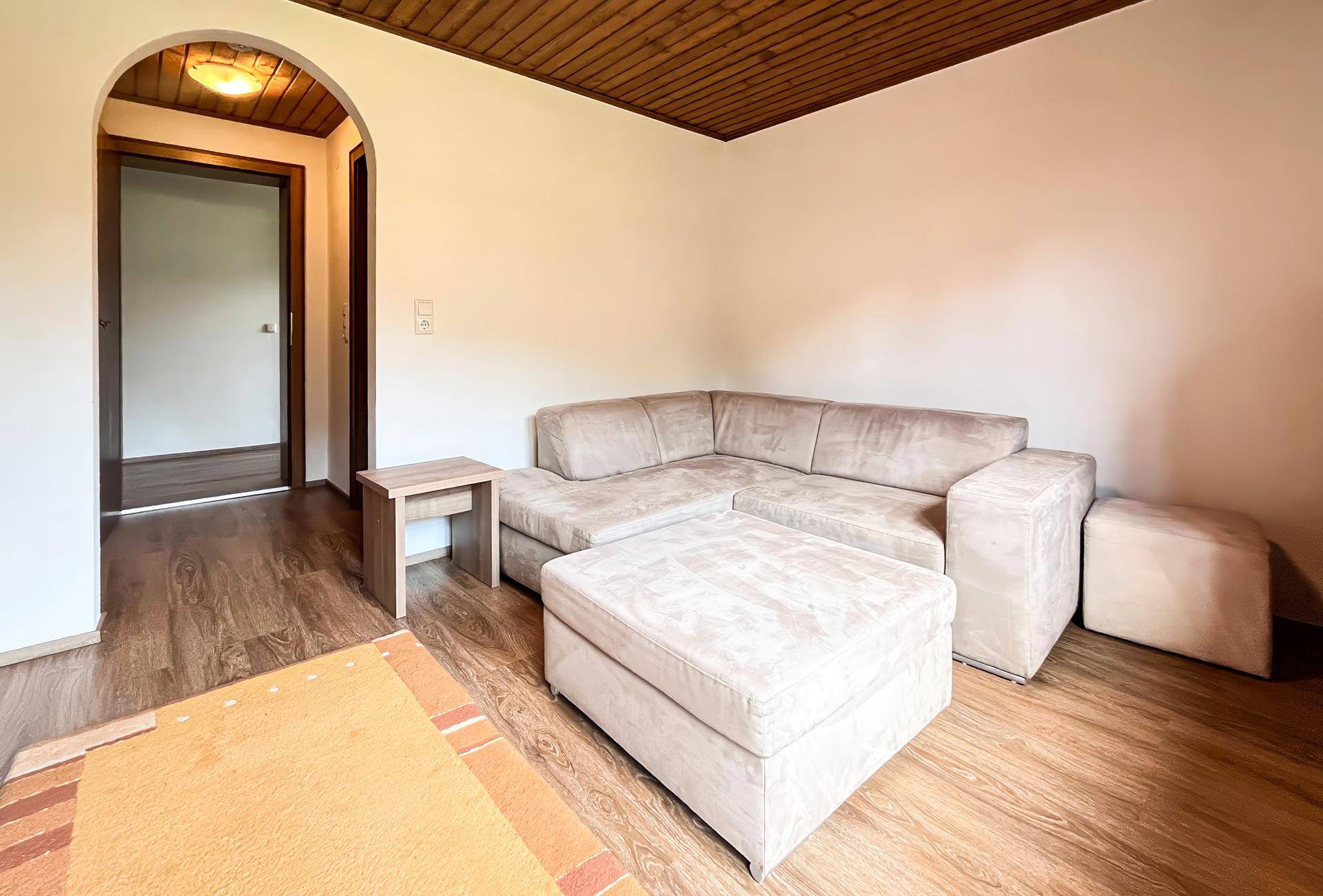 Wohnzimmer mit beigem Ecksofa, passendem Hocker und Laminatboden unter Holzdecke.