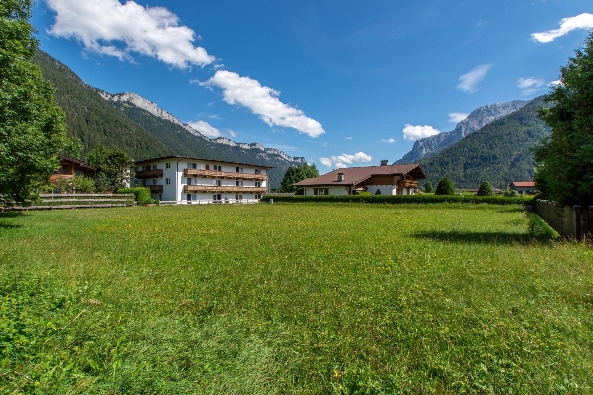 Weitläufige grüne Wiese mit mehreren Häusern und majestätischen Bergen im Hintergrund an einem sonnigen Tag.