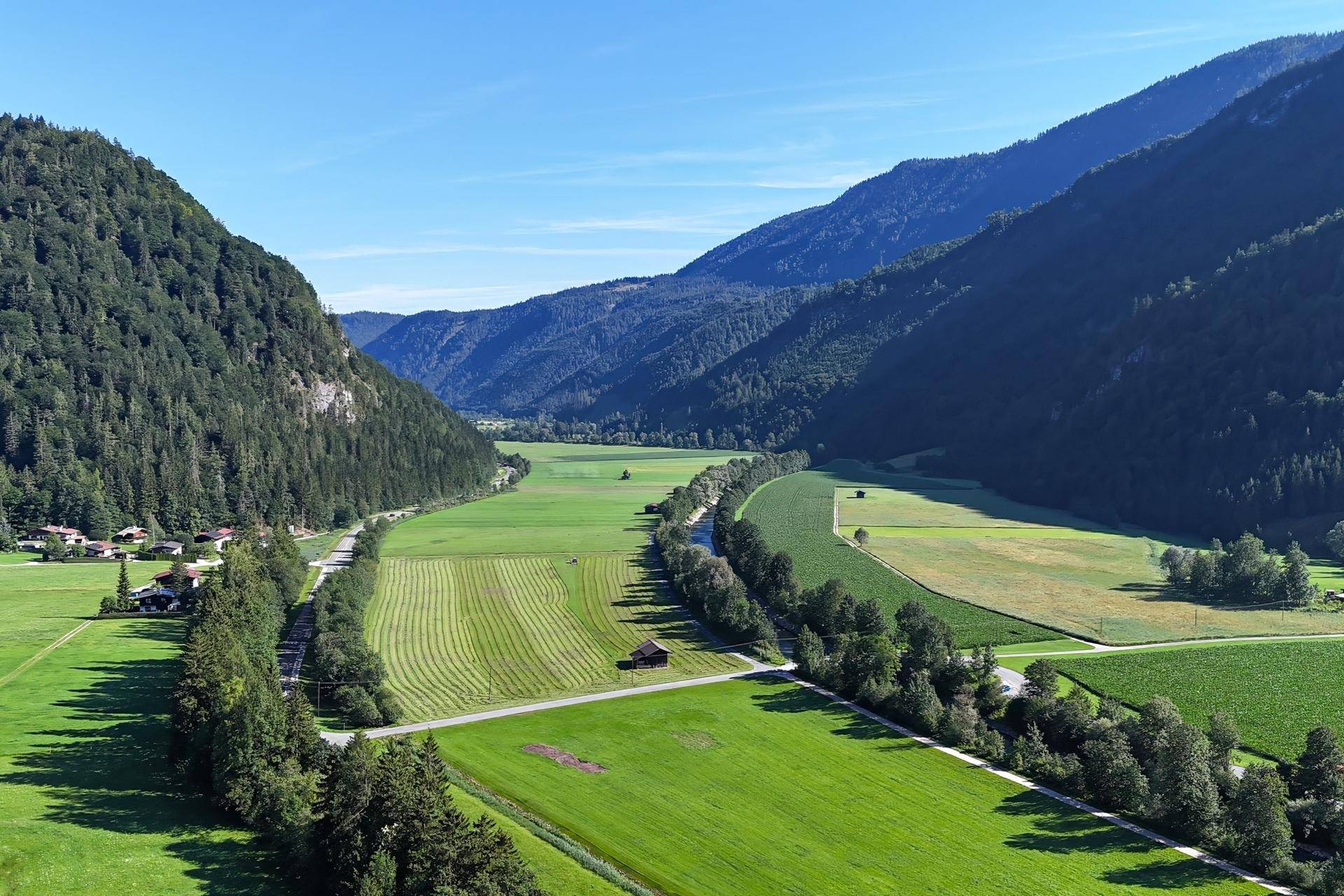 Weitläufiges Tal mit grünen Wiesen, bewaldeten Bergen und einem Fluss unter blauem Himmel.