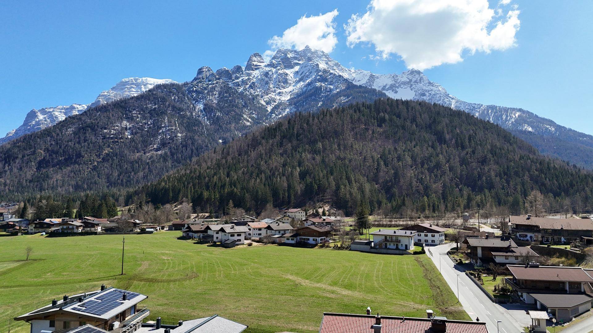 Panoramablick auf das Dorf mit schneebedeckten Bergen und grünen Wiesen unter blauem Himmel.