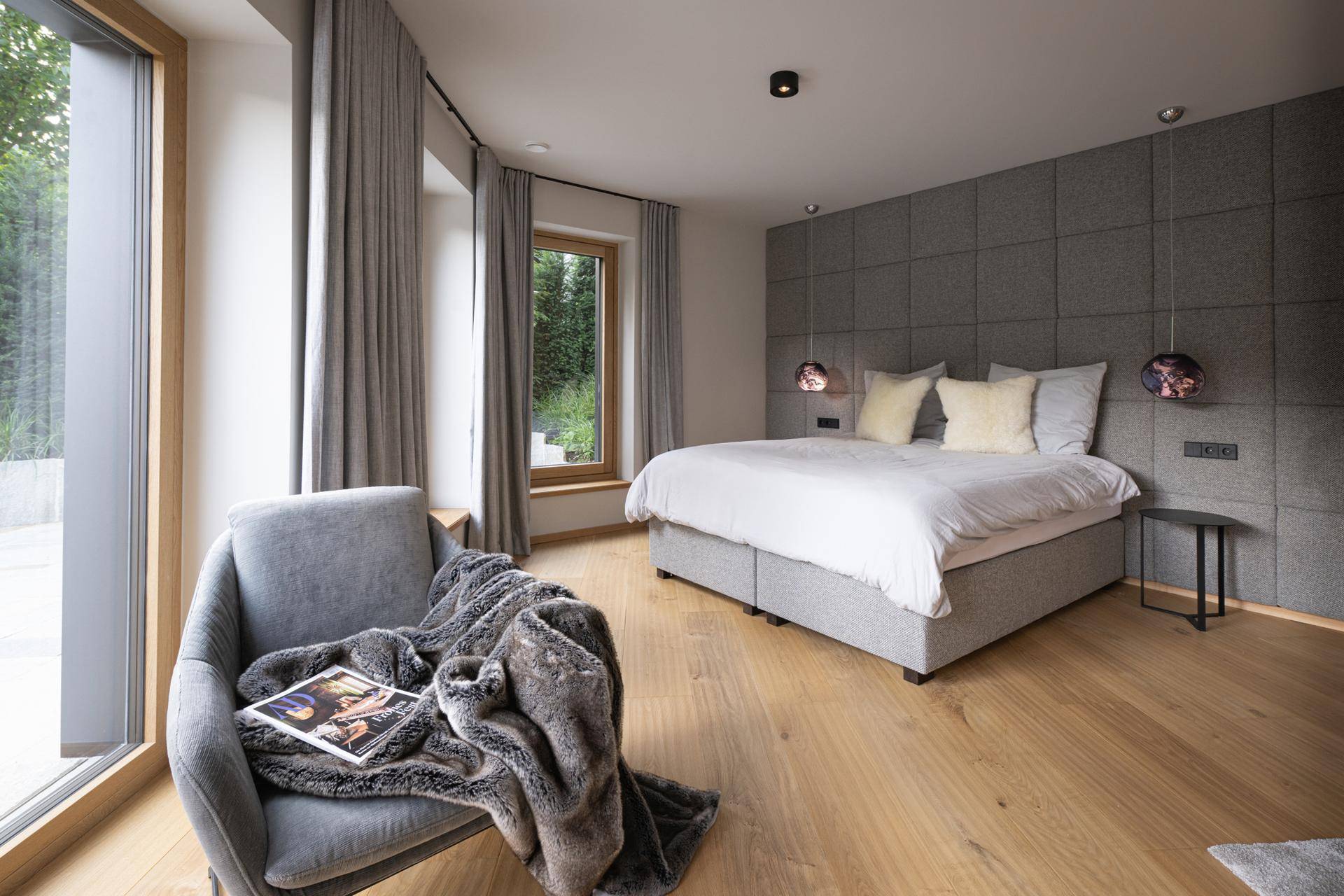 Helles Schlafzimmer mit grauem Bett, Holzboden und großen Fenstern mit Blick ins Grüne.
