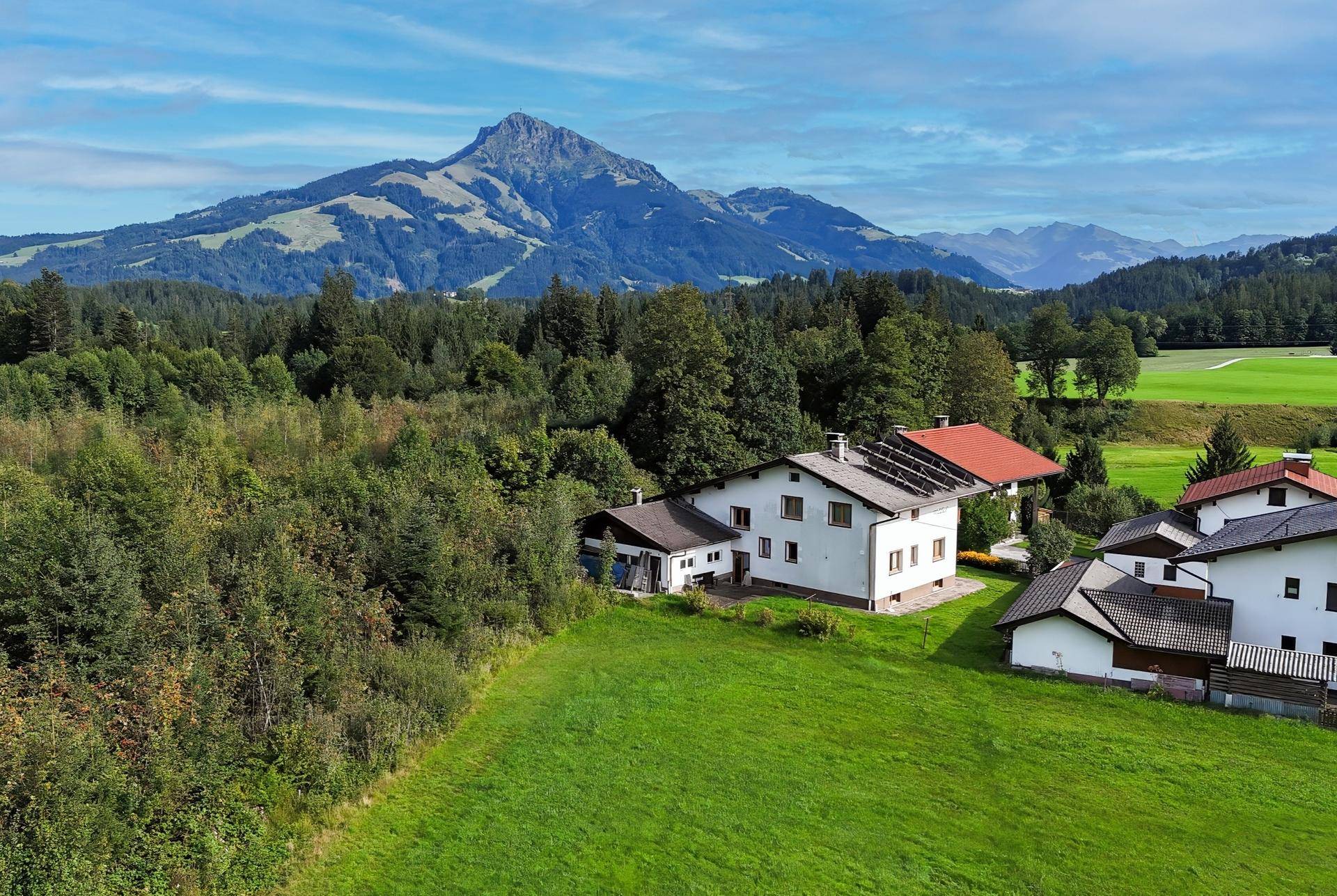 Luftaufnahme eines Hauses mit Solaranlage auf dem Dach, umgeben von grüner Natur und Bergen.