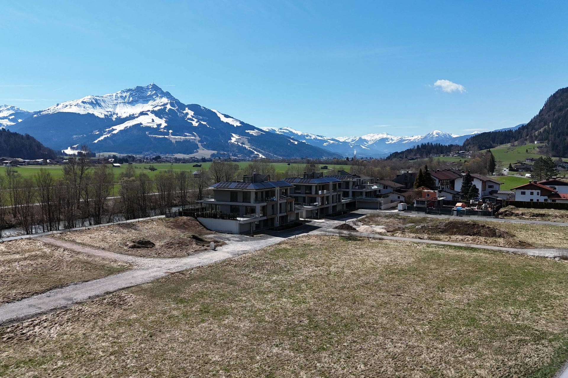 Luftaufnahme von Neubauten in alpiner Landschaft mit weiten Feldern und schneebedeckten Gipfeln.