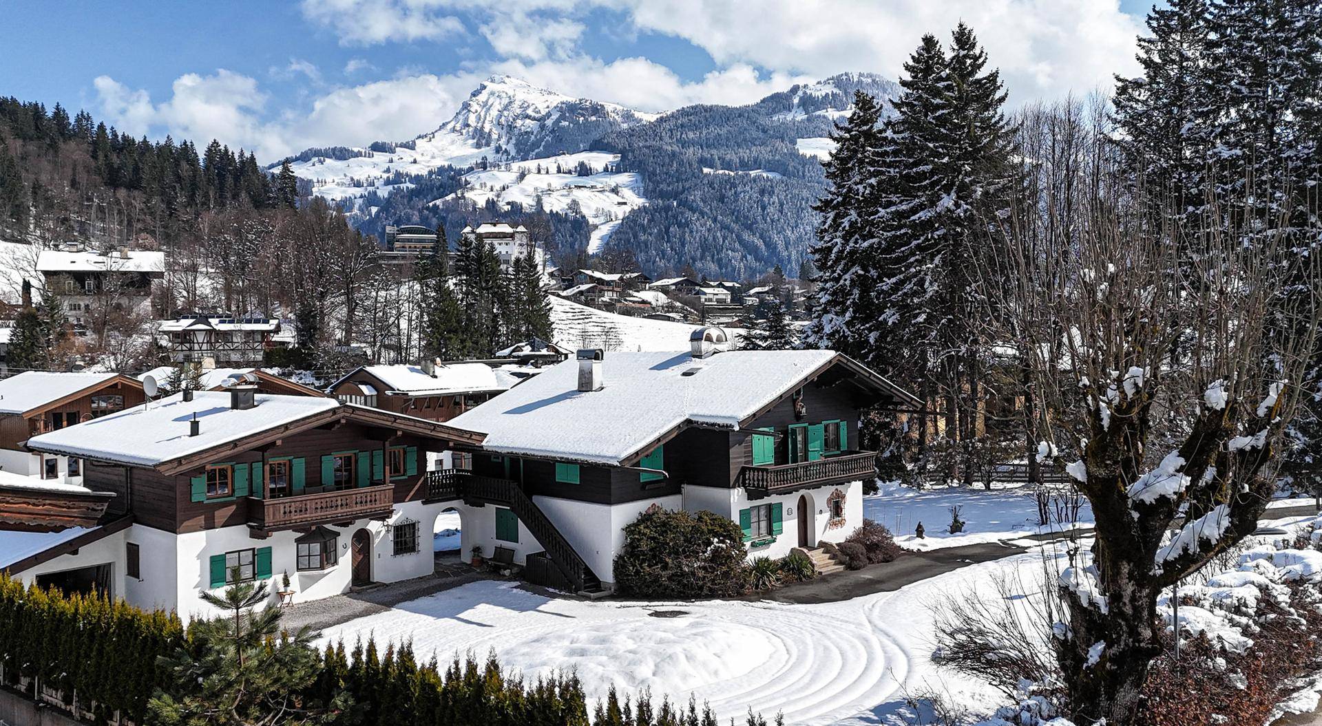 Zwei malerische Alpenhäuser im Winter, bedeckt mit Schnee, vor einer beeindruckenden Kulisse schneebedeckter Berge unter blauem Himmel.
