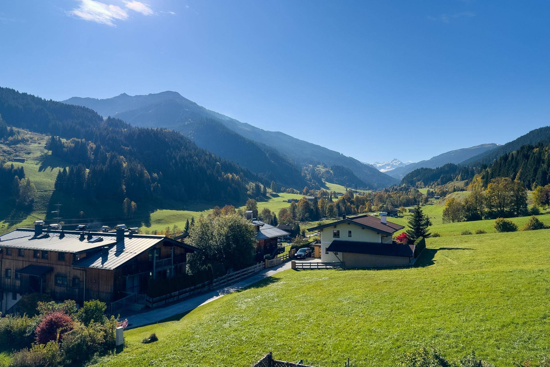 Malerische Alpenlandschaft mit grünen Hügeln, traditionellen Häusern und schneebedeckten Gipfeln in der Ferne.