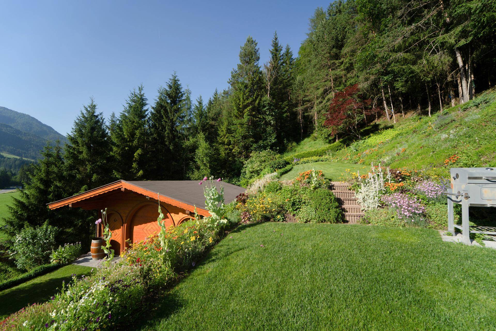 Idyllischer Gartenbereich mit blühenden Beeten, Rasenfläche und einem Holzschuppen am Waldrand.