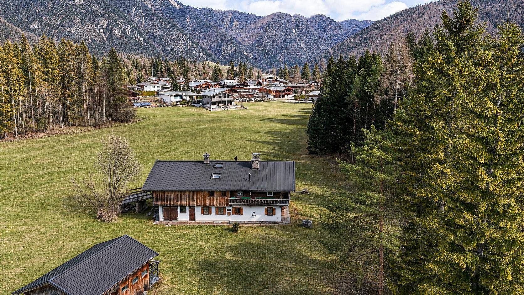 Traditionelles Haus in alpiner Umgebung mit weiten Wiesen und bewaldeten Bergen unter leicht bewölktem Himmel.