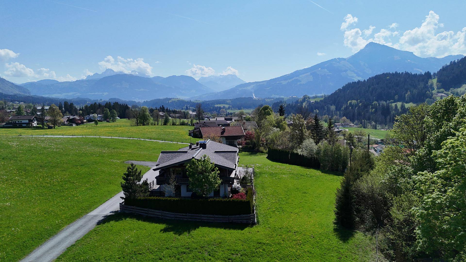 Anwesen in einer weiten grünen Landschaft mit Bergen im Hintergrund unter blauem Himmel.