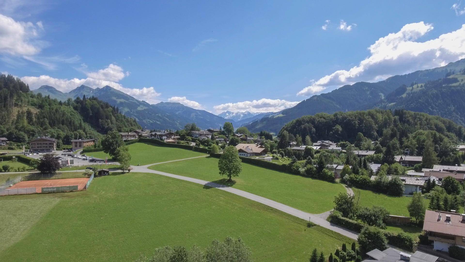 Luftaufnahme einer ländlichen Siedlung in einem grünen Tal, umgeben von bewaldeten Bergen unter blauem Himmel.