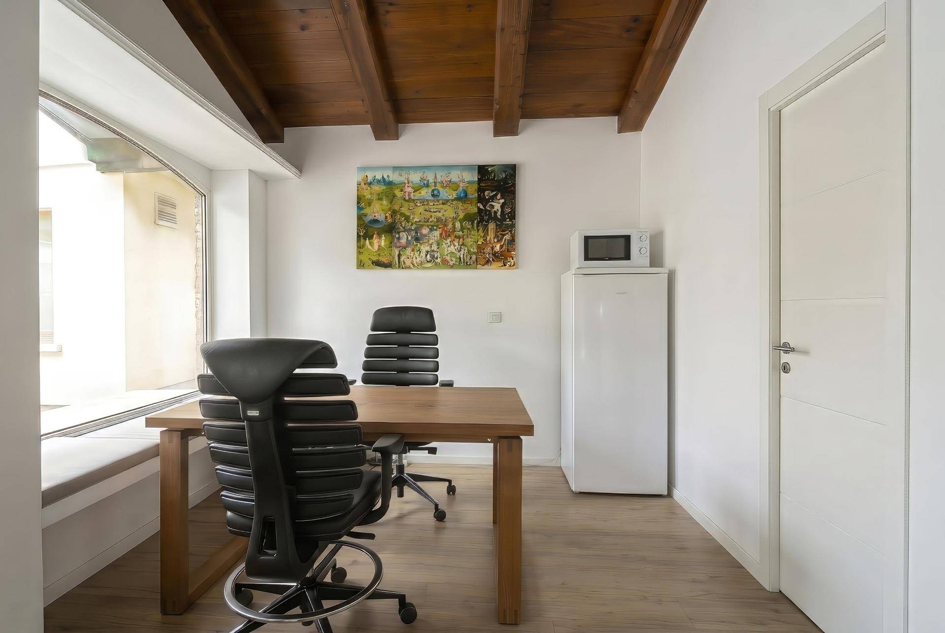Funktionaler Bürobereich mit Holztisch, Bürostühlen, Kühlschrank und Mikrowelle unter Holzbalkendecke.