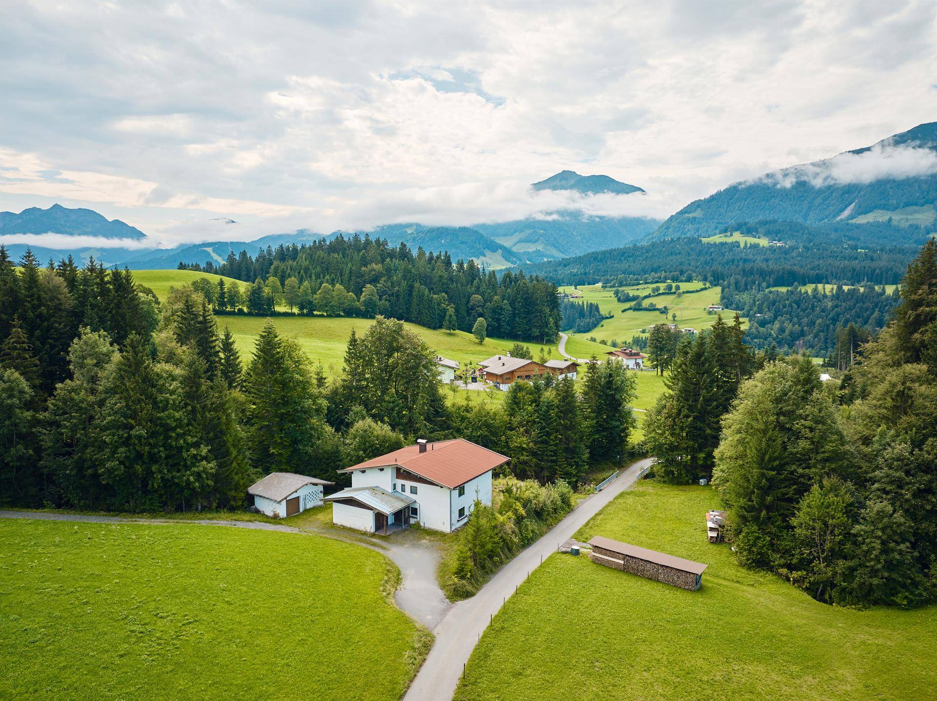 Luftaufnahme eines Einfamilienhauses mit rotem Dach, umgeben von grünen Wiesen und Wäldern in den Bergen.