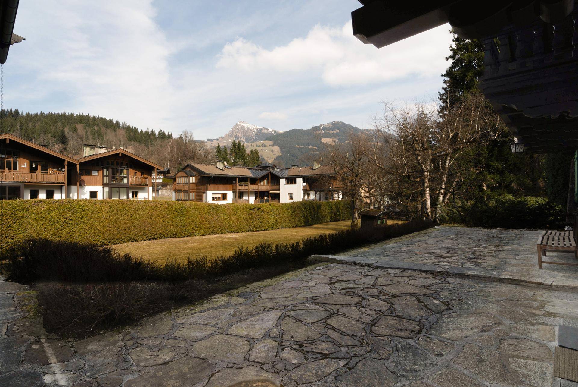 Geräumige Steinterrasse mit Holzbank und Blick auf die umliegende Landschaft und die Berge, umgeben von einer grünen Hecke.