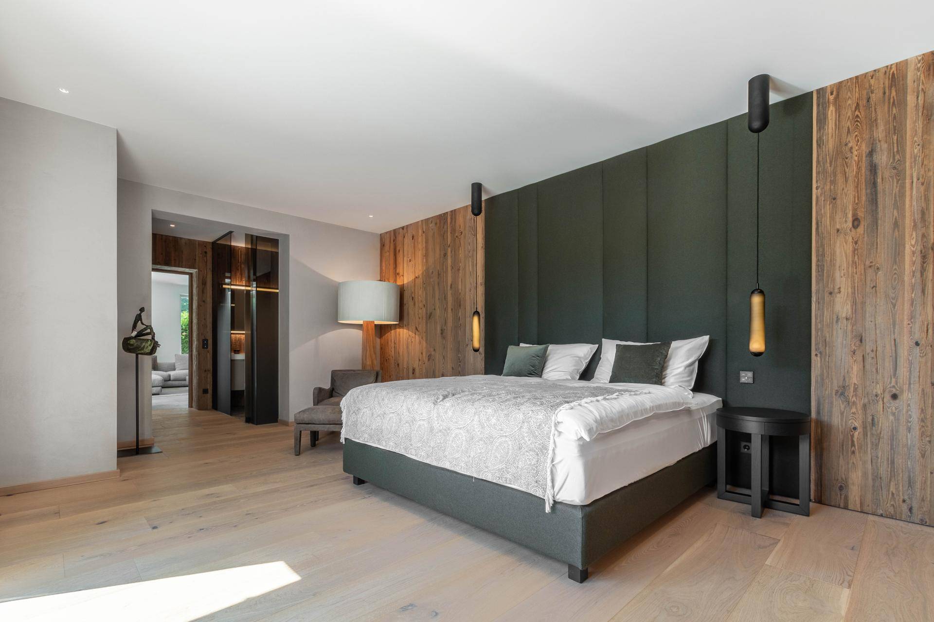 Elegantes Schlafzimmer mit Holzwand, gepolstertem Kopfteil und hellem Holzboden, mit Zugang zu einem weiteren Raum.