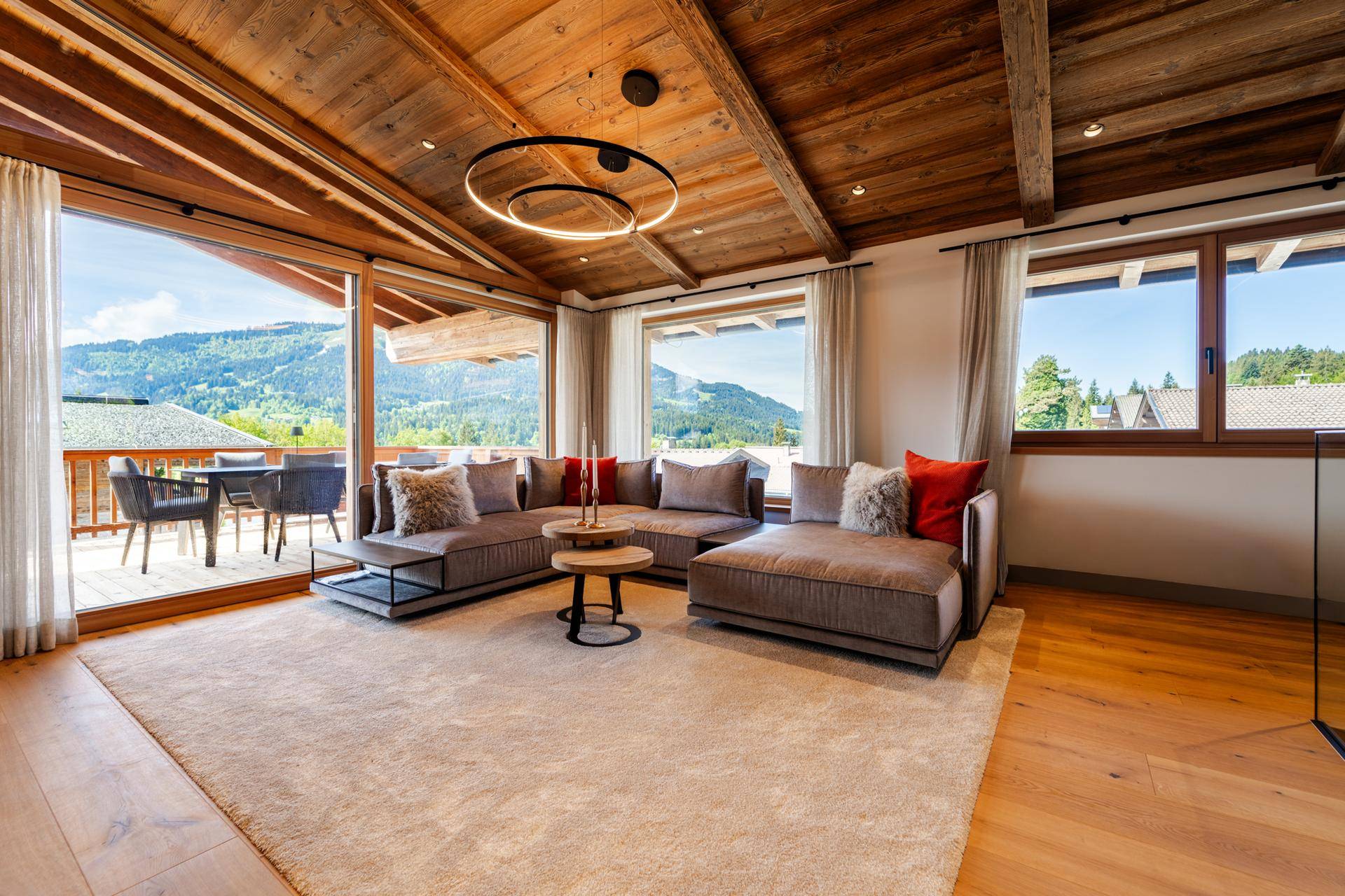 Helles Wohnzimmer mit großem Sofa, Holzbalkendecke und Zugang zu einem sonnigen Balkon mit Bergblick.