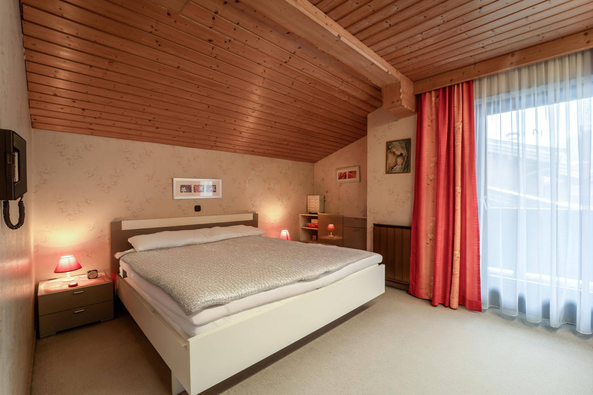 Schlafzimmer mit Holzdecke, Teppichboden und Zugang zu einem kleinen Balkon.