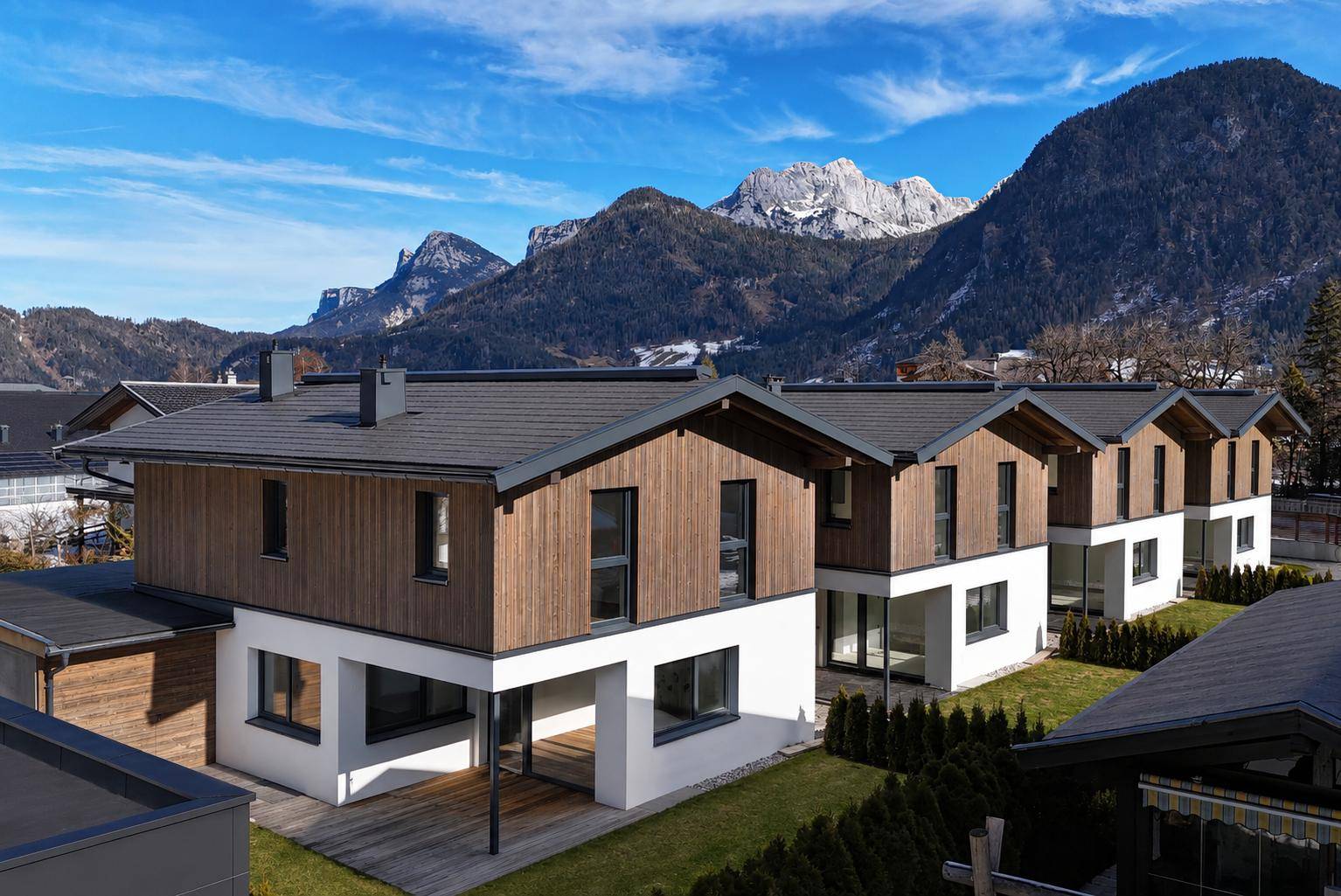 Moderne Reihenhäuser mit Holzfassade und Satteldach vor einer beeindruckenden Bergkulisse.