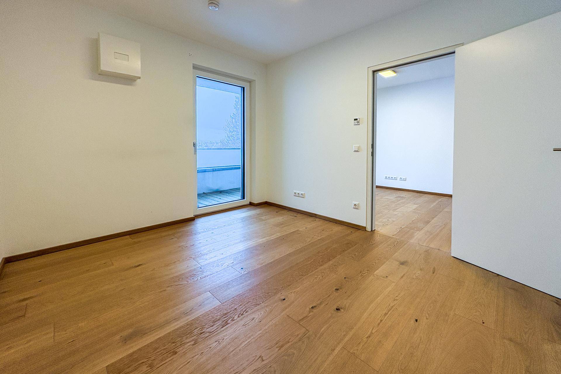 Leeres Zimmer mit Holzboden, weiß gestrichenen Wänden und Zugang zu einem Balkon.