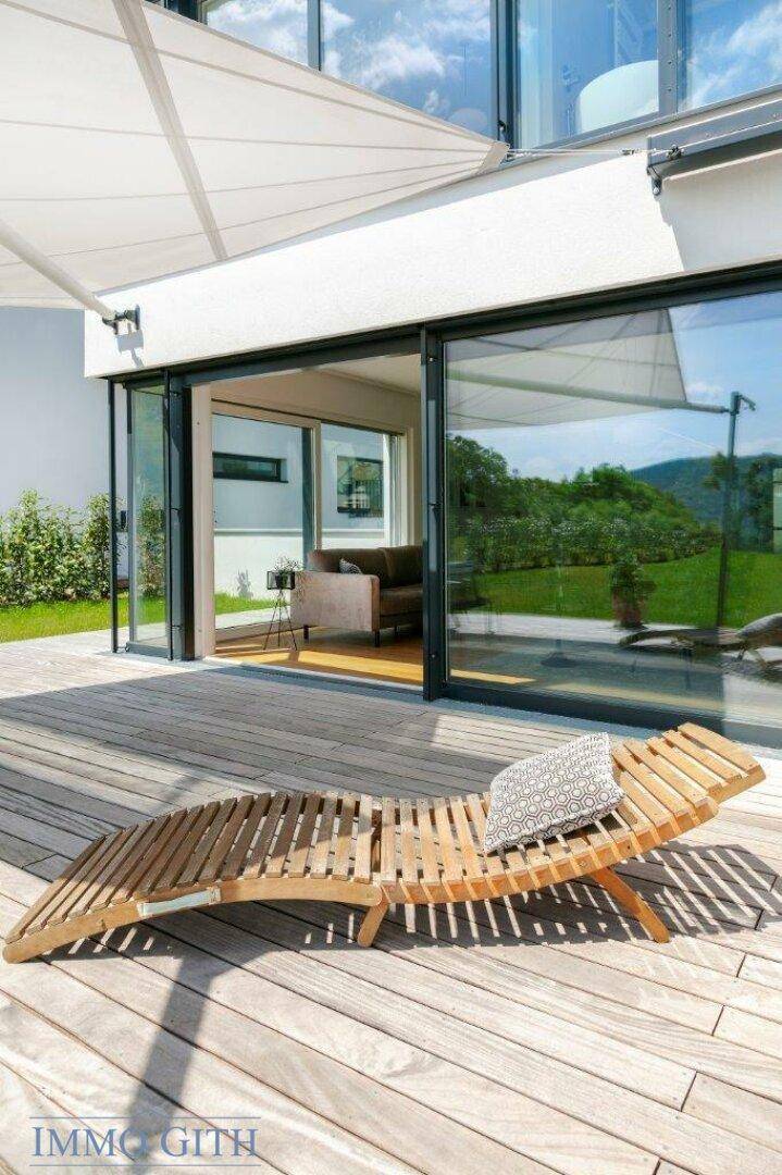 Holzliege auf der Terrasse mit Blick auf den grünen Garten und die großen Fenster des Hauses.