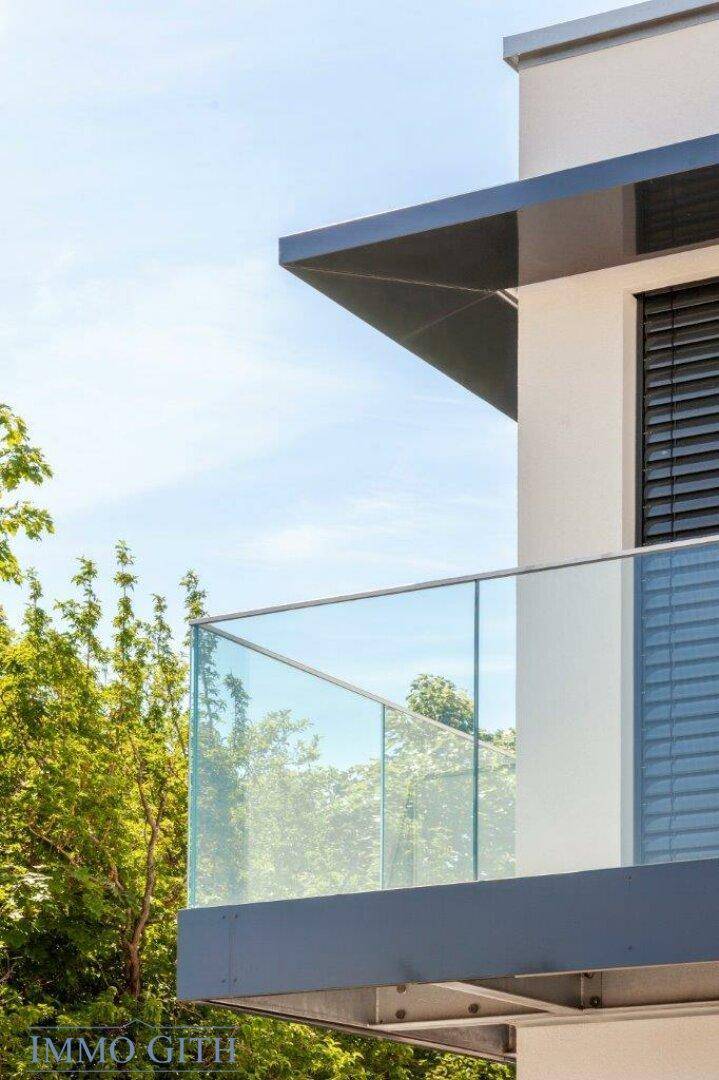 Moderner Balkon mit Glasbrüstung und Außenjalousien, umgeben von Bäumen.