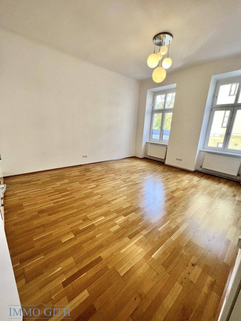 Helles, leeres Zimmer mit Parkettboden und zwei großen Fenstern, bereit zur individuellen Gestaltung.