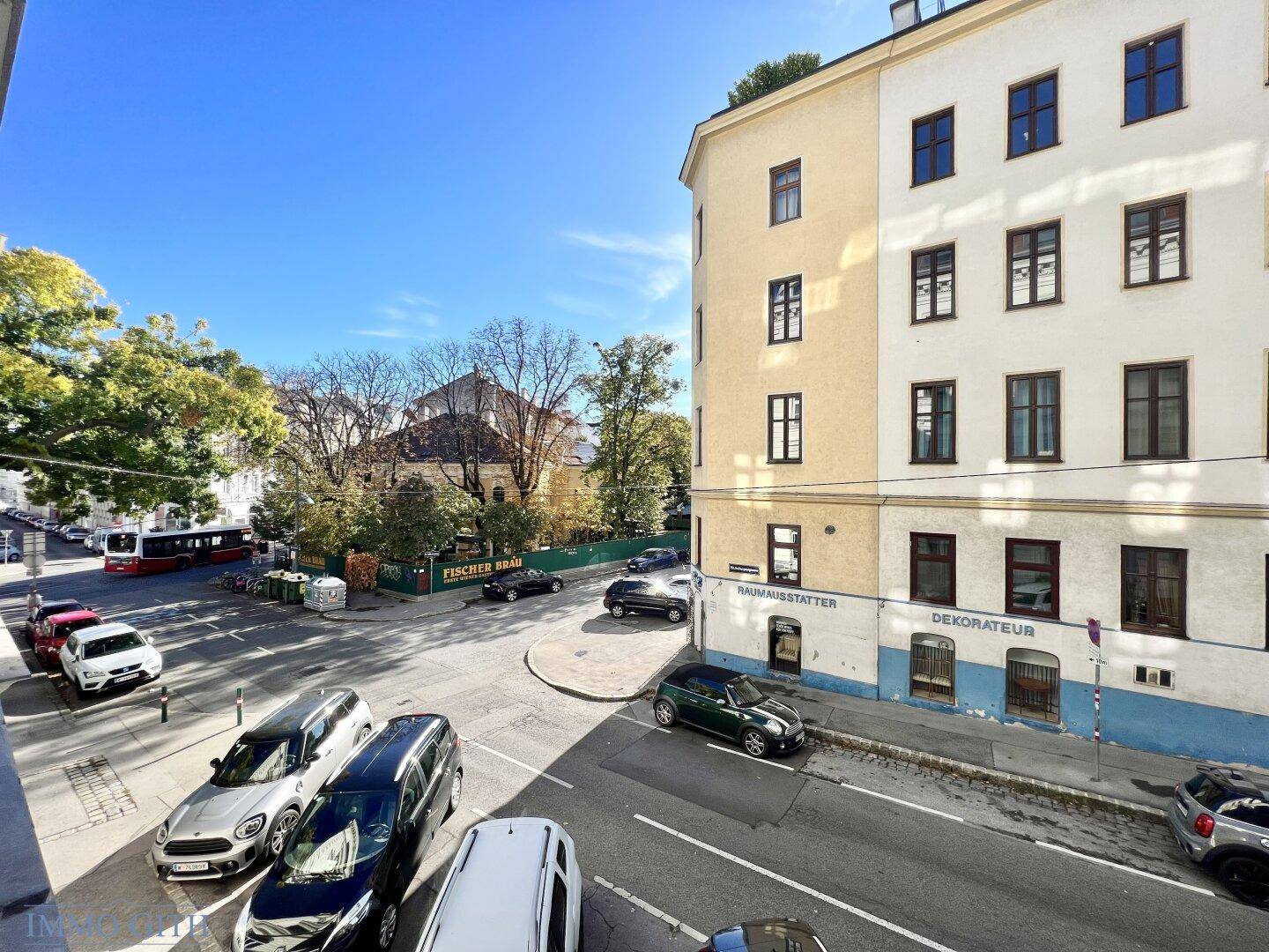 Panoramablick auf eine städtische Kreuzung mit Bäumen und Gebäuden unter blauem Himmel.