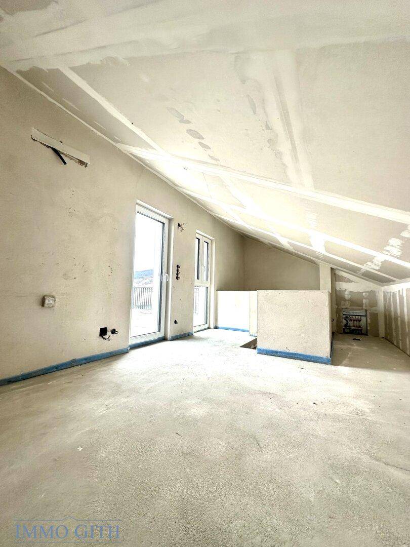 Dachgeschosszimmer im Rohbau mit schrägen Wänden, Fenstertüren und Blick auf die umliegende Landschaft.