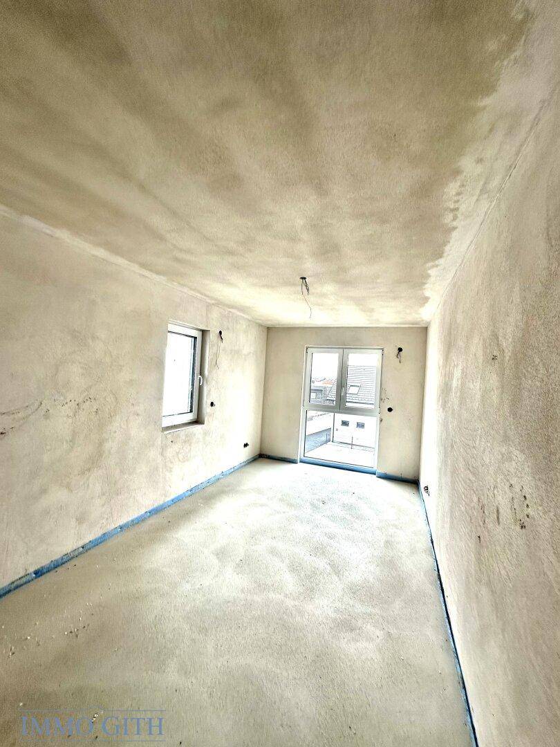 Rohbauzimmer mit zwei Fenstern, die viel Tageslicht hereinlassen, und unfertigen Wänden und Decken.