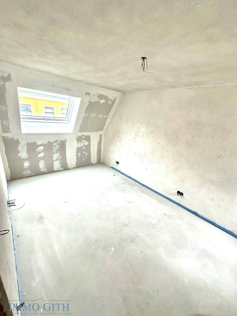 Dachgeschosszimmer im Rohbau mit Dachfenster, schrägen Wänden und viel Tageslicht für eine helle Atmosphäre.