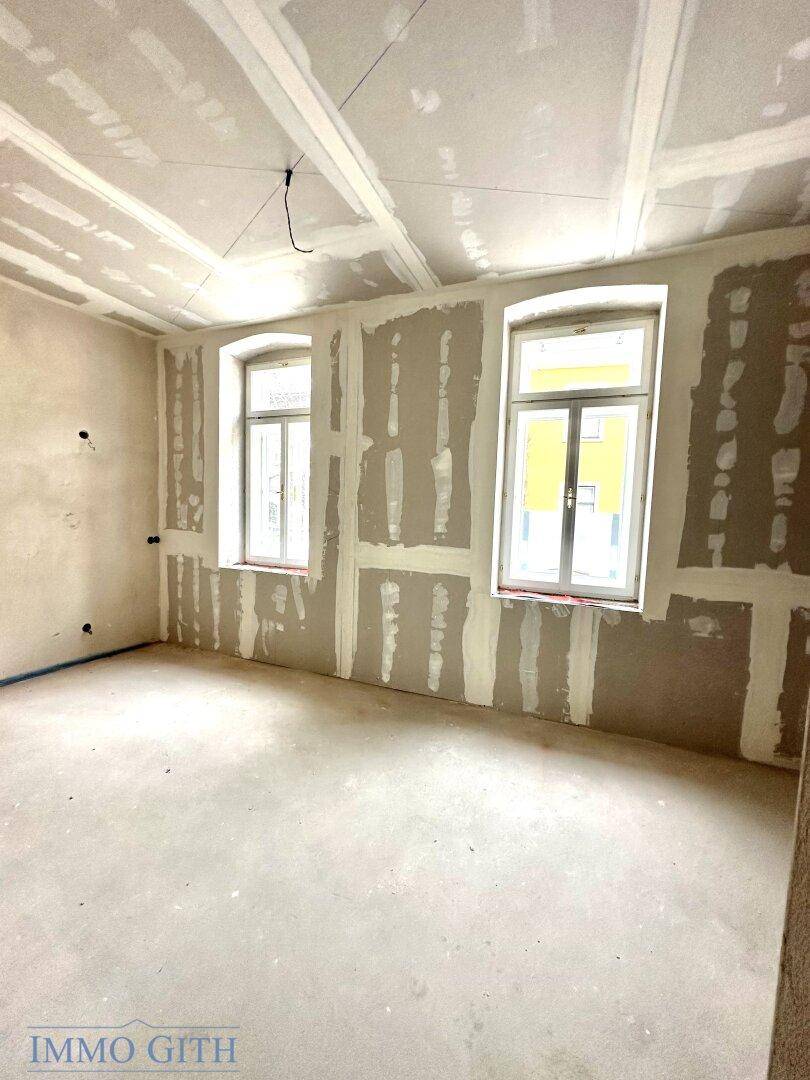 Raum im Rohbau mit zwei Fenstern, unfertigen Wänden und Decken, bereit für den Innenausbau.