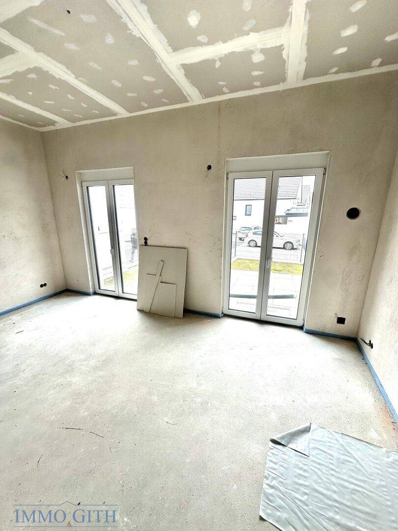 Unfertiger Raum mit großen Fenstertüren, die viel Licht hereinlassen, und vorbereiteten Wänden und Decken.