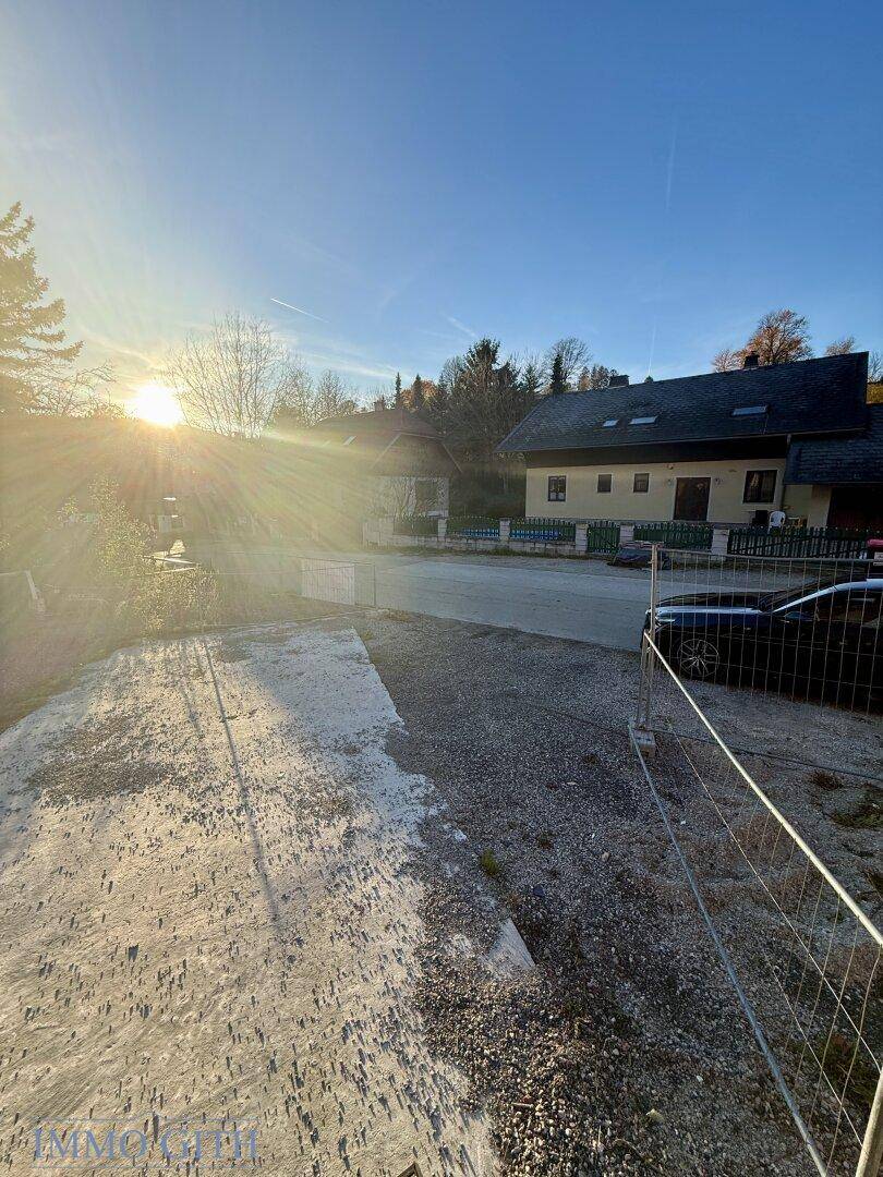 Blick über einen Schotterhof auf eine ländliche Straße und weitere Häuser im Sonnenlicht.