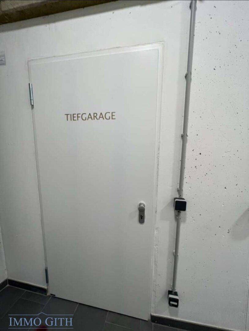 Weiße Tür mit der Aufschrift "Tiefgarage" in einem schlichten Flur mit sichtbaren Leitungen.