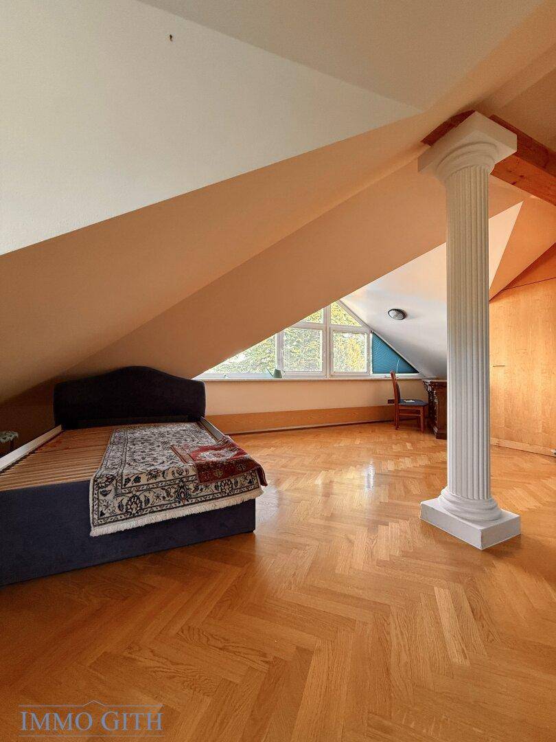 Helles Dachzimmer mit Fischgrätparkett, Bett und dekorativer Säule unter der Dachschräge.