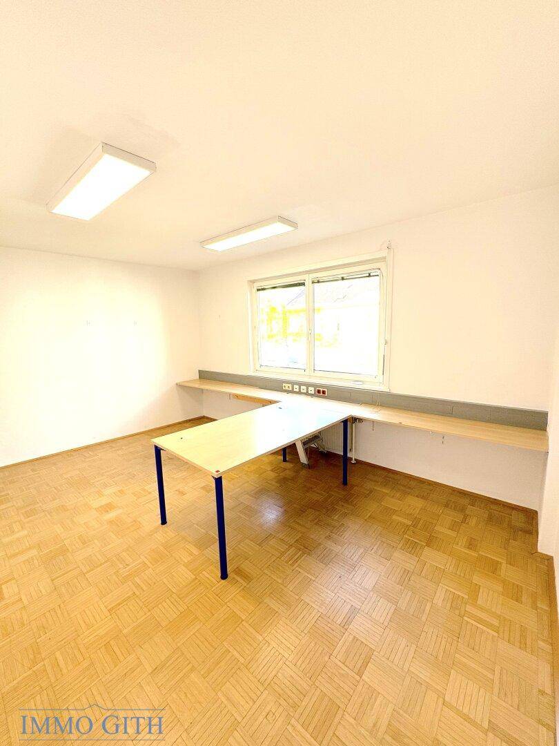 Einfacher Büroraum mit Holzboden, Fenster und langen Arbeitsflächen entlang der Wand.