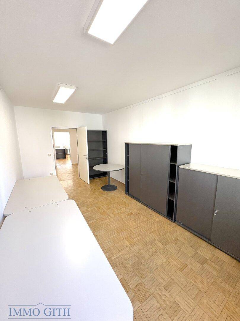 Geräumiger Büroraum mit Holzboden, grauen Schränken und Blick in den Flur.