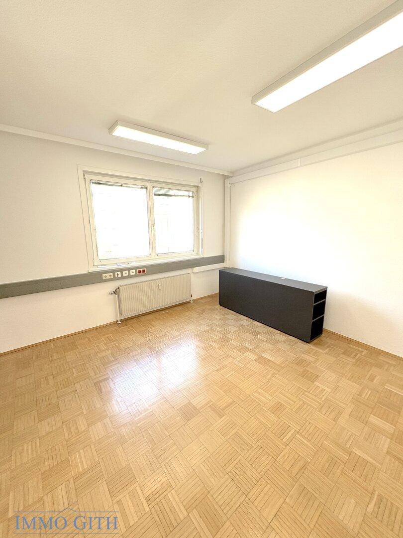 Leerer Büroraum mit Holzboden, Fenster und einem dunklen Regal, bereit zur Einrichtung.