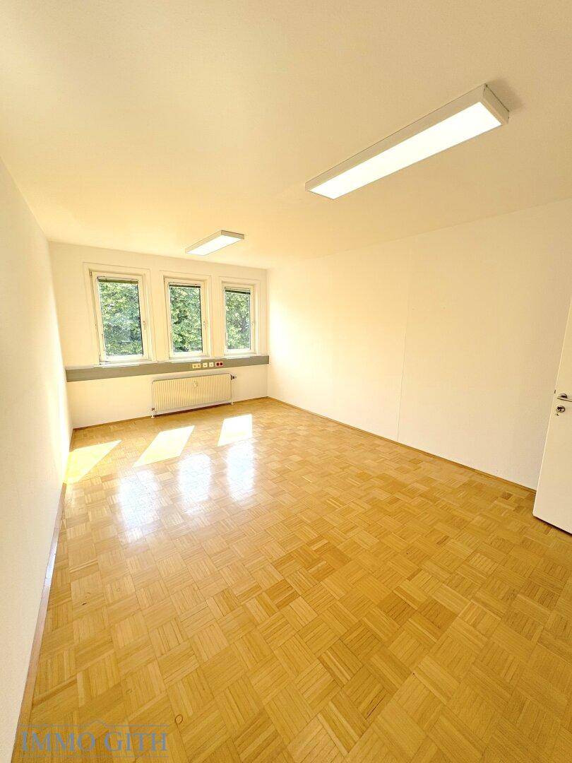 Heller Büroraum mit Holzboden und drei Fenstern, bietet viel Tageslicht und Blick ins Grüne.