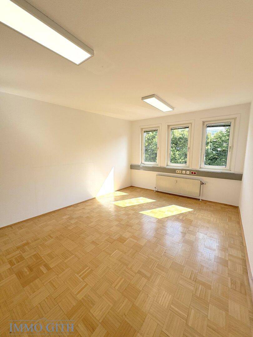 Sonniger Büroraum mit Holzboden und drei Fenstern, durchflutet von natürlichem Licht.