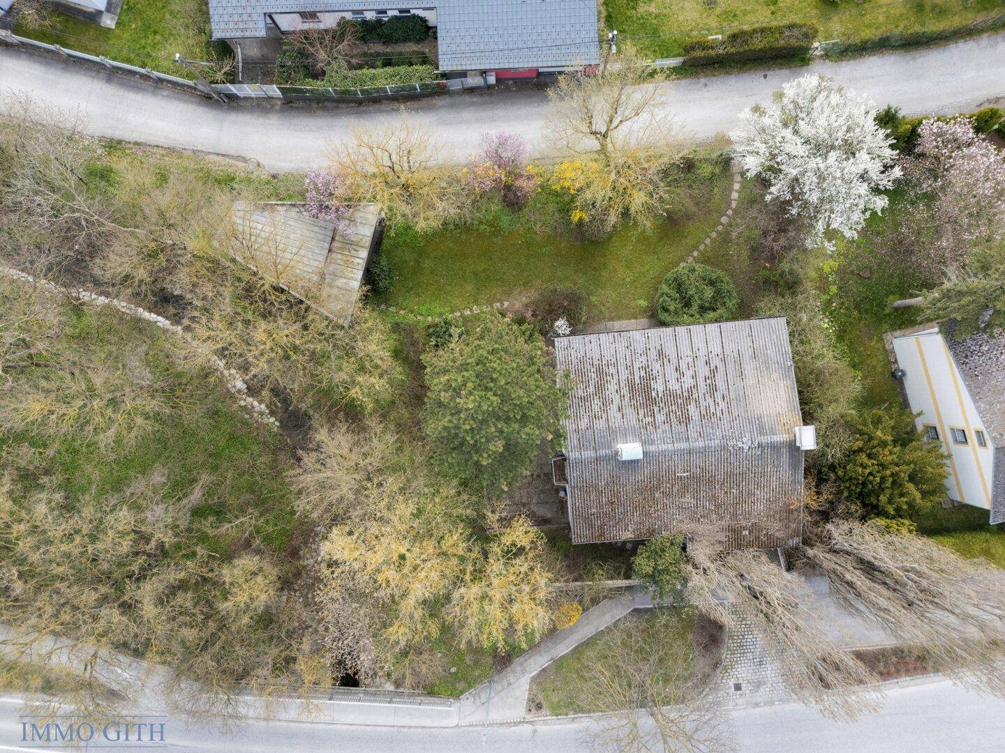 Draufsicht des Grundstücks mit Haus, Garten, Wegen und umliegender Vegetation.