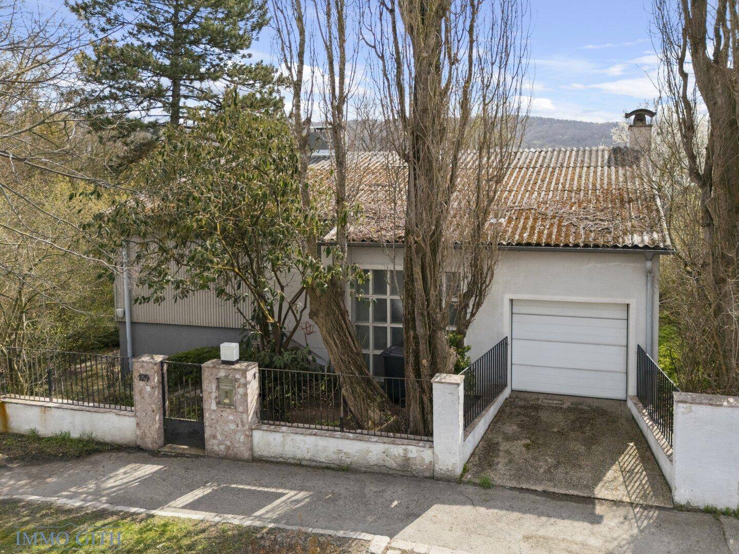 Ansicht der Hausfassade mit Garage, Einfahrt und umgebender Vegetation an einem sonnigen Tag.