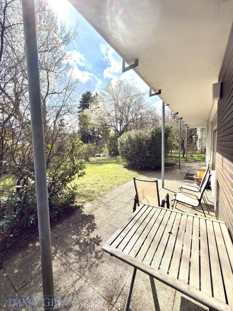 Überdachte Terrasse mit Holztisch und Stühlen, umgeben von Grünflächen und Bäumen.