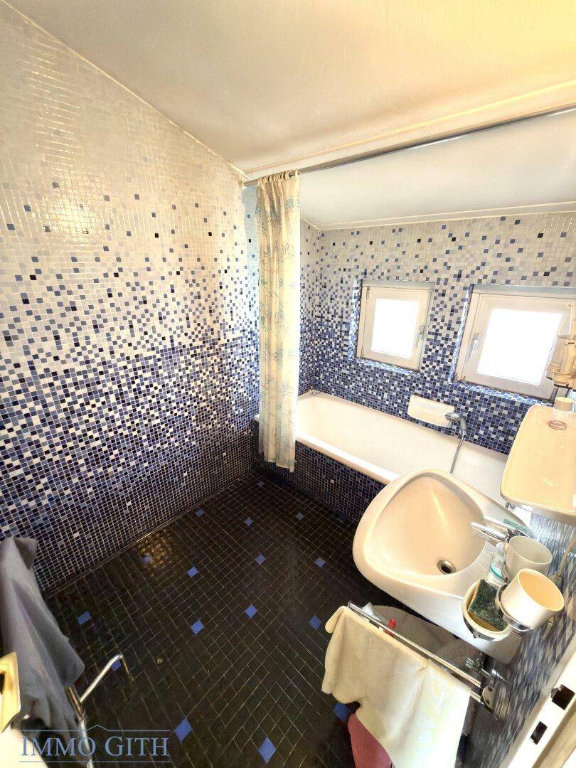 Badezimmer mit blauen Mosaikfliesen, Badewanne, Waschbecken und zwei kleinen Fenstern.