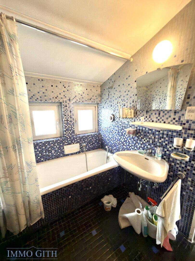 Helles Badezimmer mit blauen Mosaikfliesen, Badewanne, Waschbecken und Spiegel.