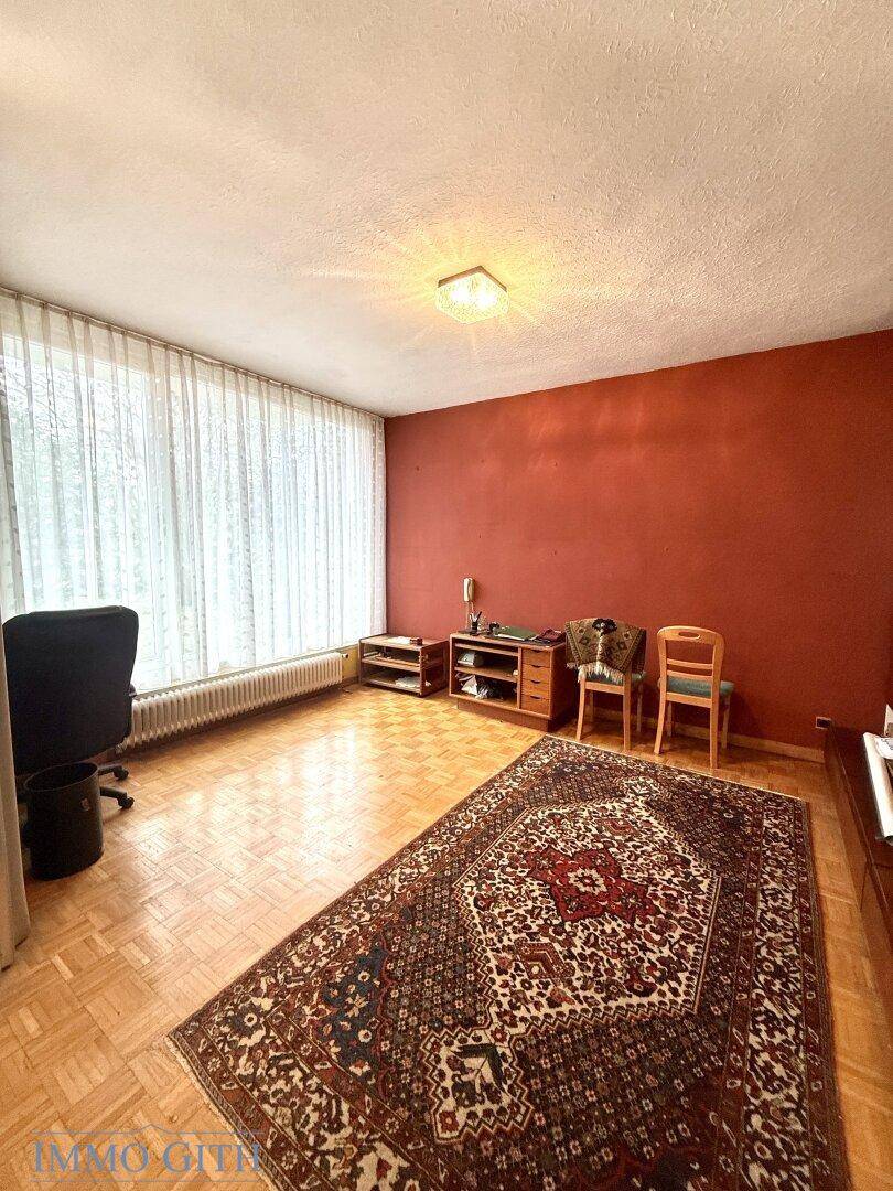 Wohnbereich mit Parkettboden, roter Akzentwand und großem Fenster mit Vorhängen.